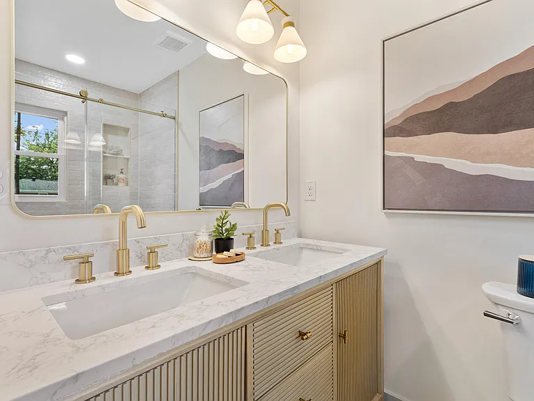 Double vanity with a fluted oak cabinet, marble counter, brass faucets, a backlit mirror, and dusty-rose mountain art