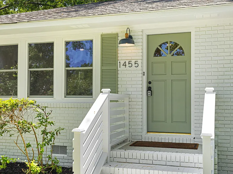 Sage-green front door set in white painted brick, with a black barn-light sconce, brass house numbers, and green louvered shutters