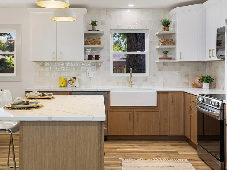 White and natural-oak kitchen with a farmhouse apron sink, gold gooseneck faucet, marble island and a sunlit window over the sink.