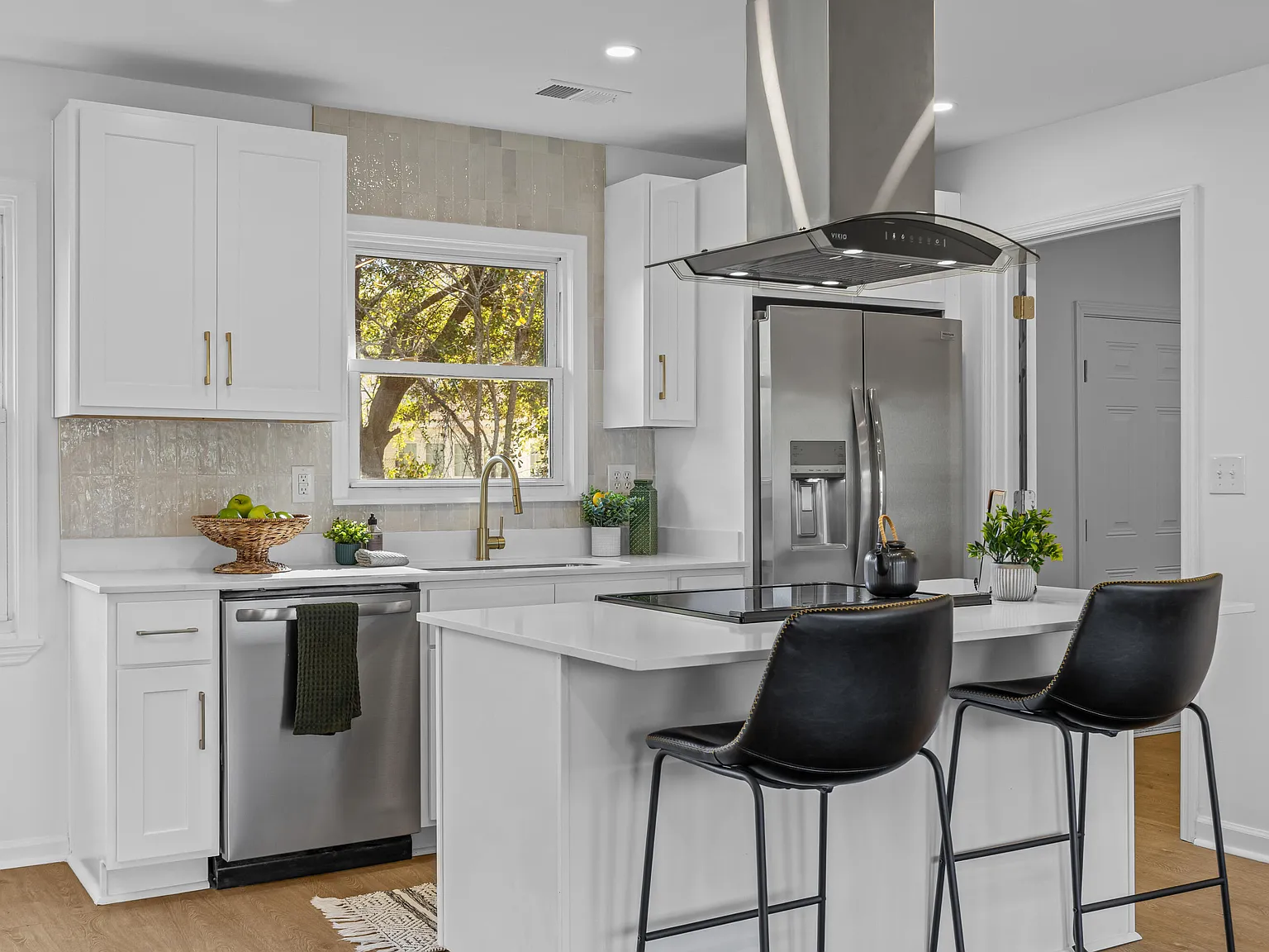 White shaker kitchen with a waterfall white-quartz island, black leather stools, a curved stainless range hood, and a gold faucet
