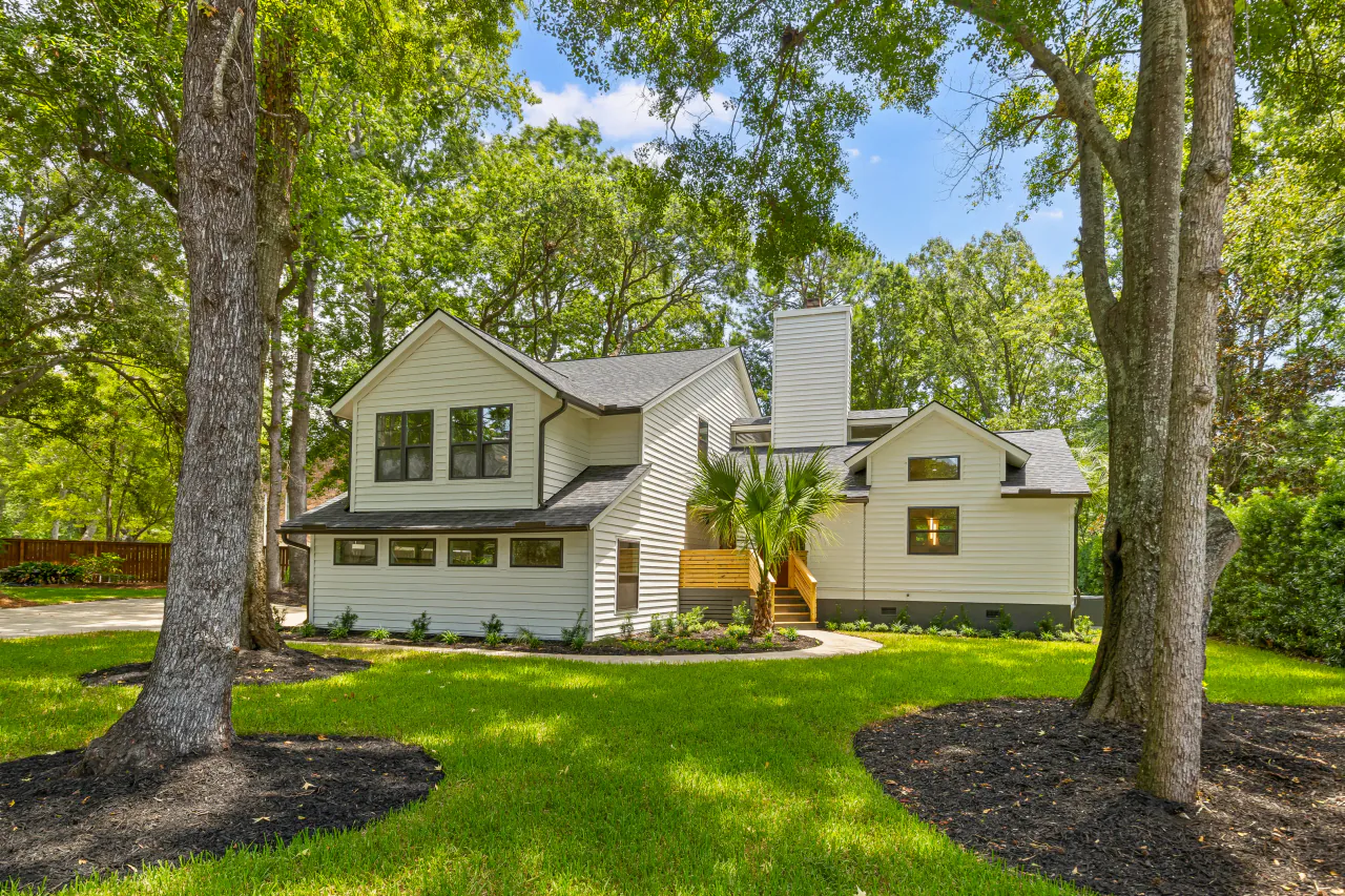 Two-story coastal farmhouse with soft blue-gray siding, framed by mature live oaks over a deep green lawn.