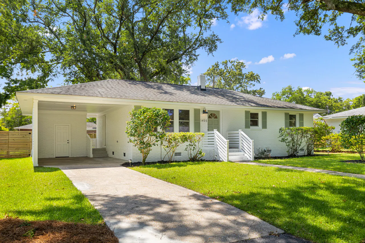 Cream-brick mid-century ranch with a carport, a clean concrete walkway, and a broad oak canopy over a green lawn.